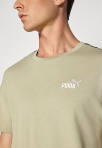 Olivgrünes T-Shirt aus weichem, glattem Stoff mit einem weißen gestickten Logo auf der Brust und einem klassischen Rundhalsausschnitt.