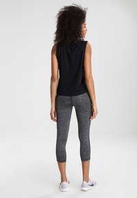 Czarny bezrękawnik o luźnym kroju, w połączeniu z szarymi, teksturowanymi legginsami za kostkę. Model ma na sobie jasnoszare buty sportowe.