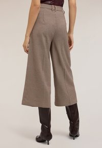 Pantalons larges à carreaux marron et beige avec une taille haute, des passants de ceinture et une finition texturée, associés à des bottes marron jusqu'aux genoux.