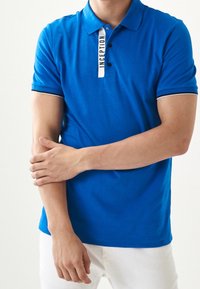 Modrá polo košile s bílým pruhovaným límcem, dvěma předními knoflíky a štítkem "INCEPTION". Tkanina vypadá hladce a pružně.