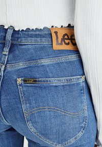 Siniset farkut, joissa on teksturoitu pinta, varustettu ruskealla nahkapaikalla, jossa on "Lee"-merkintä, sekä pienellä mustalla tarralla takataskun lähellä.