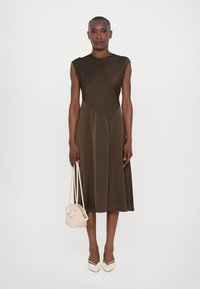 Rochie A-line de culoare maro, cu un guler geometric, confecționată dintr-un material neted. Modelul ține în mână o poșetă mică din țesătură bej. Pantofii cu toc ascuțit de culoare ivorie completează look-ul.