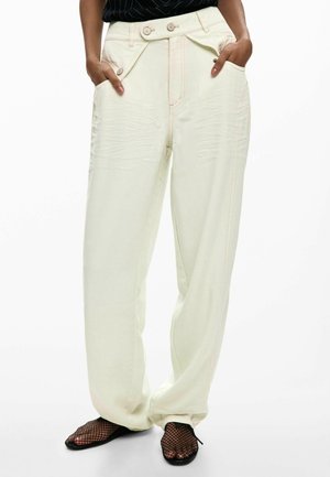 Persona che indossa pantaloni a vita alta, a gamba larga, di colore bianco sporco con bottoni e tasche, abbinati a scarpe nere in rete a rete. Mani nelle tasche.