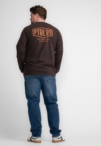 Bruine lange mouwen t-shirt met oranje tekst op de achterkant, gecombineerd met lichtblauwe jeans en witte sneakers, gezien vanuit de achterkant.