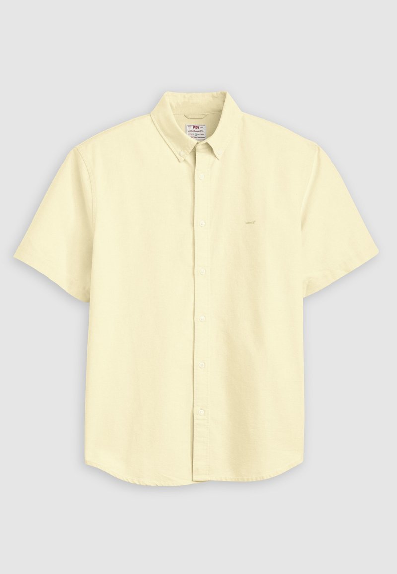 Camisa de manga corta en amarillo pálido con botones, cuello puntiagudo y logo sutil en el pecho izquierdo, mostrada sobre un fondo gris.