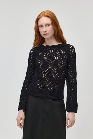 Rothaarige Frau, die einen schwarzen, langärmeligen, offen gestrickten Pullover und einen schwarzen Rock trägt, vor einem schlichten hellen Hintergrund steht.