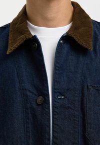 Gros plan sur une personne portant une veste en denim sombre avec un col en velours côtelé marron, par-dessus un t-shirt blanc à col rond.