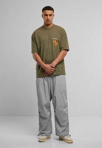 Olivengrøn t-shirt med korte ærmer og et logo på brystet, kombineret med løstsiddende lysegrå bukser og beige sneakers.