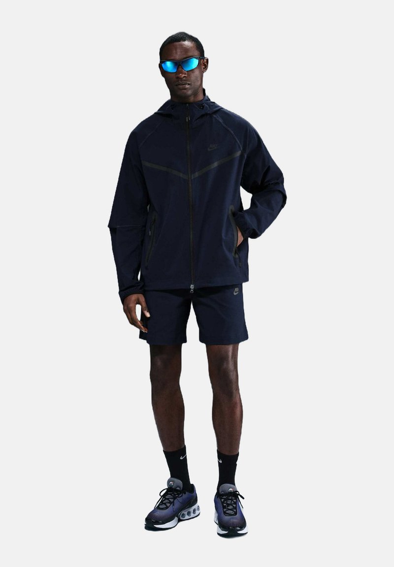 Nike mörkblå vindjacka med dragkedja fram, kontrasterande sömmar och matchande shorts. Tillbehören inkluderar blå solglasögon och sportskor.