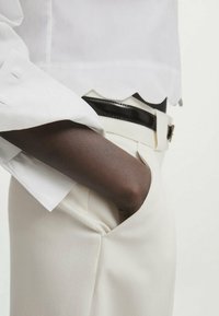 Chemise blanche à bords festonnés avec manches longues, associée à un pantalon crème et une ceinture noire. La main repose dans la poche, mettant en valeur la texture du tissu.