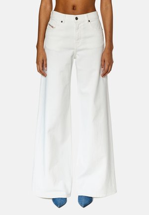 Femme portant un pantalon blanc à taille haute et jambes larges, associé à des chaussures pointues bleues, debout devant un fond clair uni.