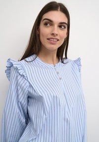 Camicia a maniche lunghe azzurro chiaro con strisce verticali bianche e marroni. Presenta dettagli arricciati sulle spalle e un collo rotondo. Tessuto dalla texture liscia.