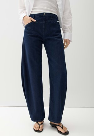 Person trägt dunkelblaue Jeans mit weitem Bein, weißes Hemd, weißes Top und schwarze Sandalen mit Absatz, eine Hand in der Tasche.