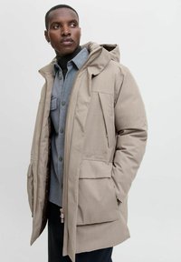 Bežna parka s kapuco, ki ima sprednji zadrgo in več žepov. Obračana je čez sivo srajco na gumbe, kar ustvarja plastičen videz.