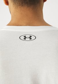 Achteraanzicht van de nek en bovenrug van een persoon die een wit overhemd draagt met een klein zwart Under Armour-logo onder de kraag.