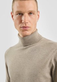 Beżowy sweter z wysokim kołnierzem wykonany z miękkiej dzianiny. Dopasowany krój z prążkowanym kołnierzem i gładką teksturą, prezentowany na neutralnym tle.