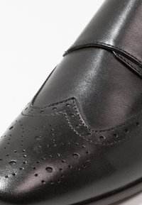 Svart lädersko med spetsig tå, perforerad detaljering och dekorativa broguedetaljer. Slät textur och elegant finish.