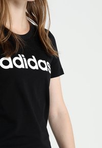 Černé bavlněné tričko s velkým bílým logem "adidas" na hrudi, krátké rukávy a klasický crew neck design.