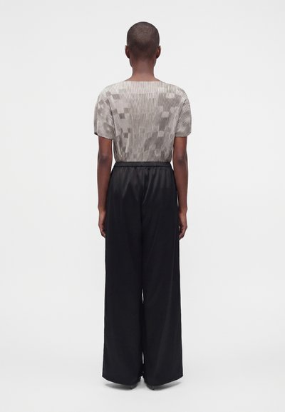 Personne debout de dos, portant un haut beige à manches courtes à motif et un pantalon large noir ample, sur un fond uni.