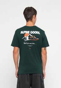 Mann som står med ryggen til, iført mørkegrønn T-skjorte med teksten "ALPINE GOODS", skikunst, fjell og orange sol på baksiden.