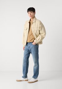 Chaqueta de mezclilla beige sobre una camisa marrón, combinada con vaqueros azules y zapatillas blancas con acentos beige. Corte slim y estilo casual.