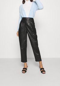 Vrouw draagt een lichtblauw zacht vest over een witte coltrui, een zwarte leren broek met hoge taille en zwarte sandalen met hak en gevlochten bandjes.