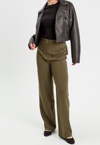 Vrouw draagt olijfgroene, wijde broek met hoge taille, zwart aansluitend topje, cropped zwarte leren jas en zwarte schoenen tegen een witte achtergrond.