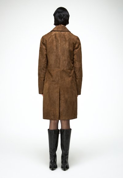 Personne aux cheveux courts noirs portant un long manteau marron et des bottes noires à talons montant jusqu'aux genoux, vue de dos sur un fond blanc.