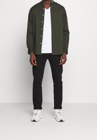 Donkergroene blouse met lange mouwen en staande kraag, witte V-hals t-shirt eronder, zwarte jeans en witte sneakers met grijze accenten.