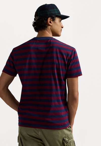 Rdeča in mornarsko modra prugasta bombažna majica s krožnim izrezom, kratkimi rokavi in sproščenim krojem, v kombinaciji z olivno zelenimi kratkimi hlačami.