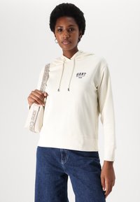 Kremowa bluza z kapturem na sznurki, z logo GANT i rokiem wydania nadrukowanym w granatowym kolorze, w połączeniu z ciemnoniebieskimi dżinsami oraz fakturowaną jasną torbą.