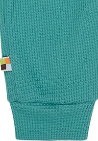 Polsino di un indumento lavorato a maglia turchese con un motivo testurizzato, dotato di un piccolo etichetta multicolore cucita sul lato.