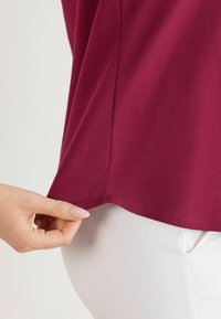 Weinrotes, strukturiertes Oberteil mit abgerundetem Saum, das sanft am Rand gezogen wird, kombiniert mit weißen Hosen. Glatter Stoff mit dezenter Detailierung.