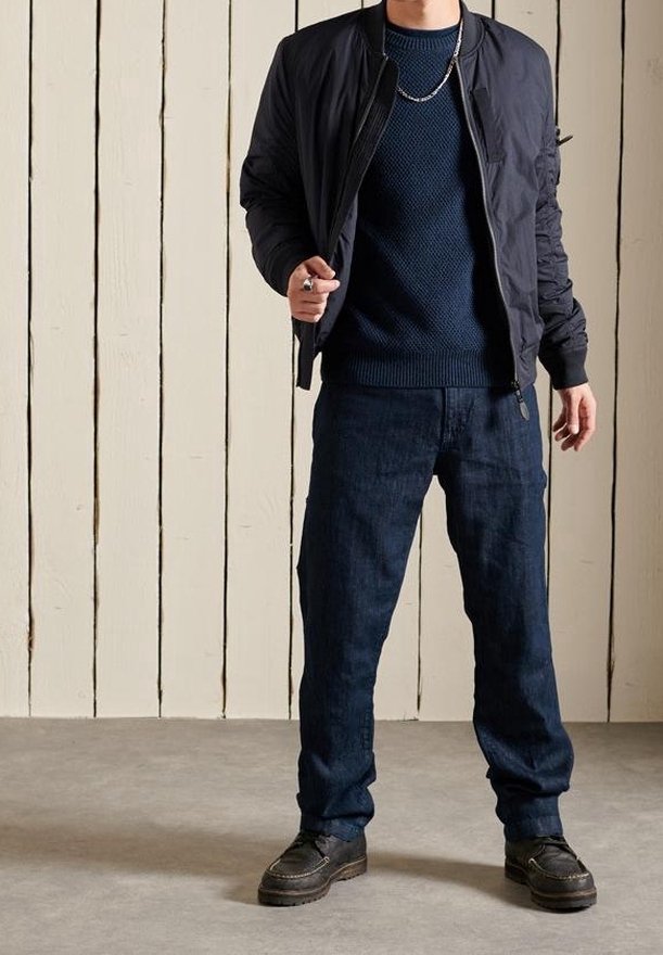 Veste bomber noire avec poignets côtelés, pull en maille marine et pantalon en denim foncé, associés à des chaussures noires, portés devant un fond texturé.