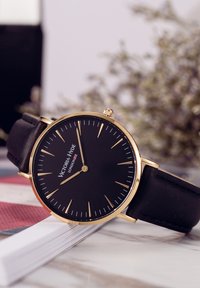 Montre noire avec un boîtier rond en or, au design minimaliste, indicateurs d'heure en or et bracelet en cuir noir. Présente le nom de la marque sur le cadran.
