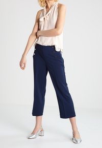 Jasnoróżowa bluzka bez rękawów z kokardą, w połączeniu z granatowymi spodniami o kroju capri z podwójnymi guzikami oraz srebrnymi sandałami na obcasie.