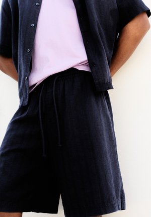 Personne portant une chemise noire à manches courtes boutonnée sur un t-shirt rose clair et un short noir avec cordon, debout devant un fond blanc.
