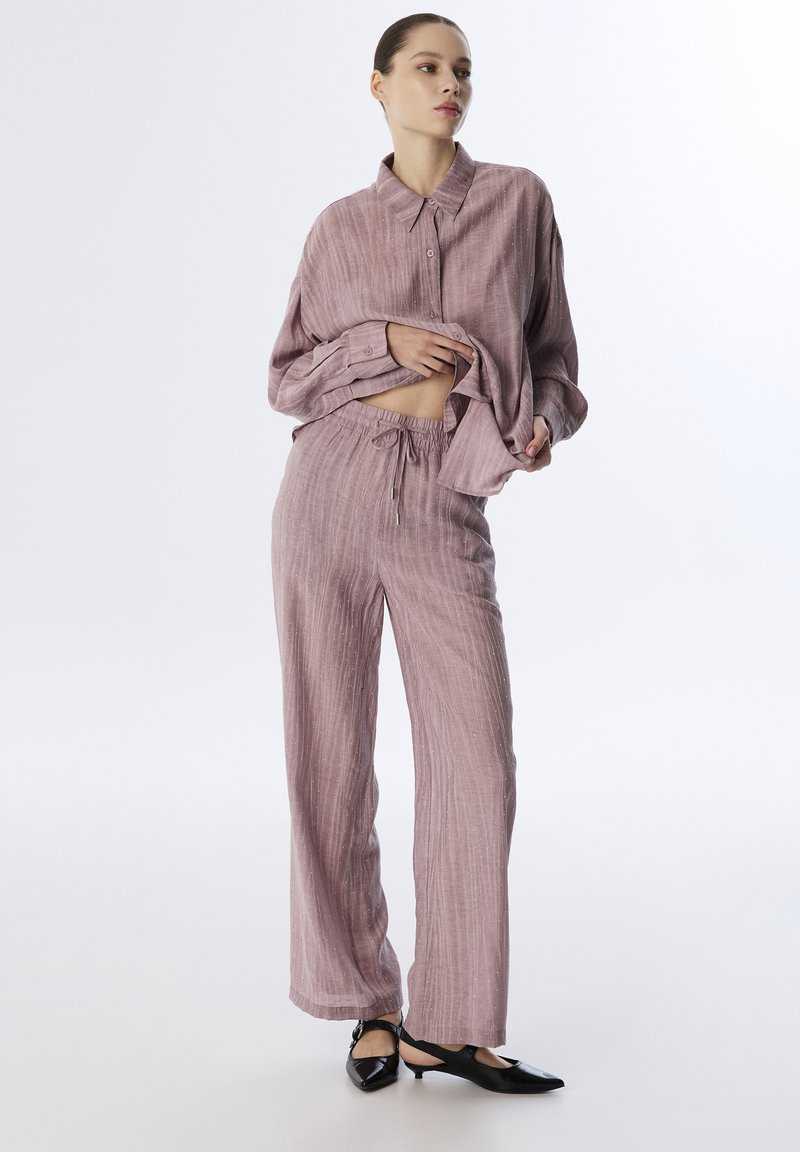 Un set da lounge di un rosa chiaro, con una camicia a maniche lunghe con bottoni morbida e pantaloni coordinati con cordoncino. Le scarpe nere a punta completano l'outfit.