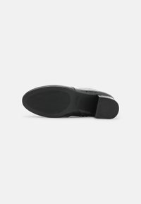 Zapato de cuero negro con una parte superior texturizada, que presenta una punta lisa y ligeramente curvada, y una suela de goma plana con ranuras horizontales.