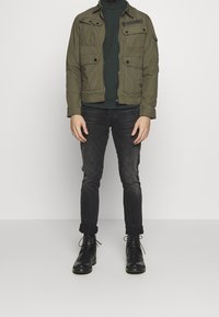 Man in een olijfgroene jas, donkergroene trui, zwarte skinny jeans en zwarte veterschoenen staand tegen een witte achtergrond.