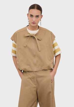 Femme aux cheveux noirs attachés porte un gilet marron clair zippé avec un nœud par-dessus une chemise à manches courtes beige et blanche à rayures et un pantalon assorti marron clair.