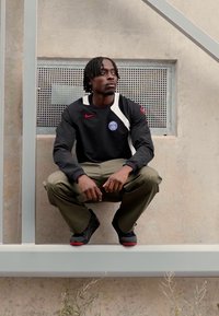 Sudadera negra de Nike con detalles y emblema blancos, pantalones caqui y zapatillas negras con suelas rojas; persona en cuclillas sobre una viga de metal.