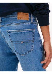 Gros plan sur une personne portant un pantalon en denim Tommy Jeans bleu clair avec une poche arrière et un écusson logo, associé à un haut à manches longues bleu marine foncé.