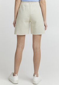 Beige bomullsshorts med elastisk midje, kne-lengde, med rå kanter. Båret med hvite joggesko, sett bakfra.