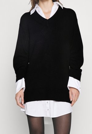 Pull en tricot noir avec un col en V et des épaules tombantes, superposé sur une chemise blanche à boutons. Poignets et ourlet côtelés. Collants noirs opaques.