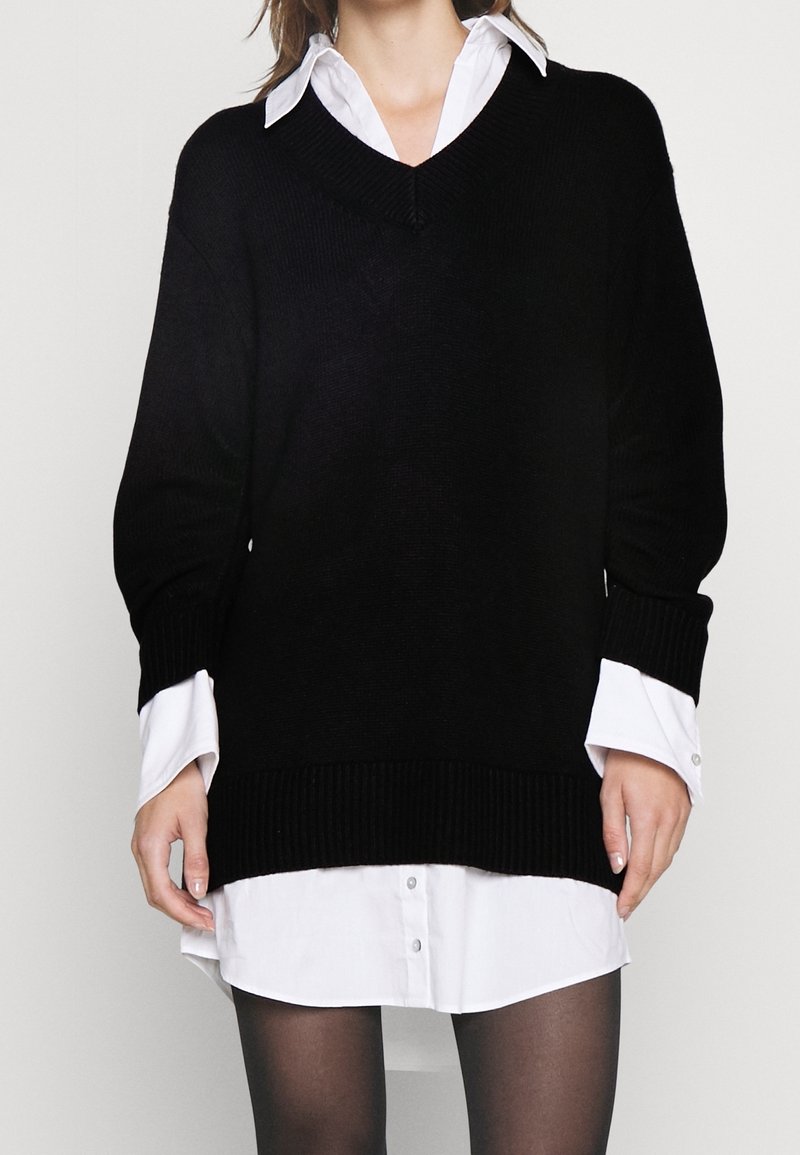 Maglione lavorato a maglia nero con scollatura a V e spalle scese, sovrapposto a una camicia bianca con bottoni. Polsini e orlo a coste. Collant neri opachi.