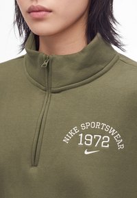 Близък план на човек, облечен в спортен топ на Nike в маслиненозелен цвят с половин цип и бял текст и лого "Nike Sportswear 1972".