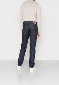 Jeans de mezclilla azul oscuro con corte recto, con costuras naranjas, dos bolsillos traseros con un diseño curvado y una etiqueta de marca en la cintura.