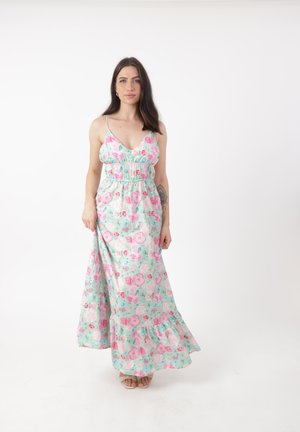 Kobieta o długich ciemnych włosach w nieslekawym pastelowym kwiecistym maksi sukience, stojąca na tle jednolitego białego tła.