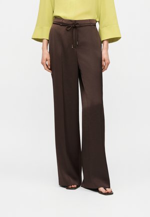 Woman wearing high-waist brown wide-leg pants with a thin belt tied in a bow, paired with a light green long-sleeve top and black sandals.